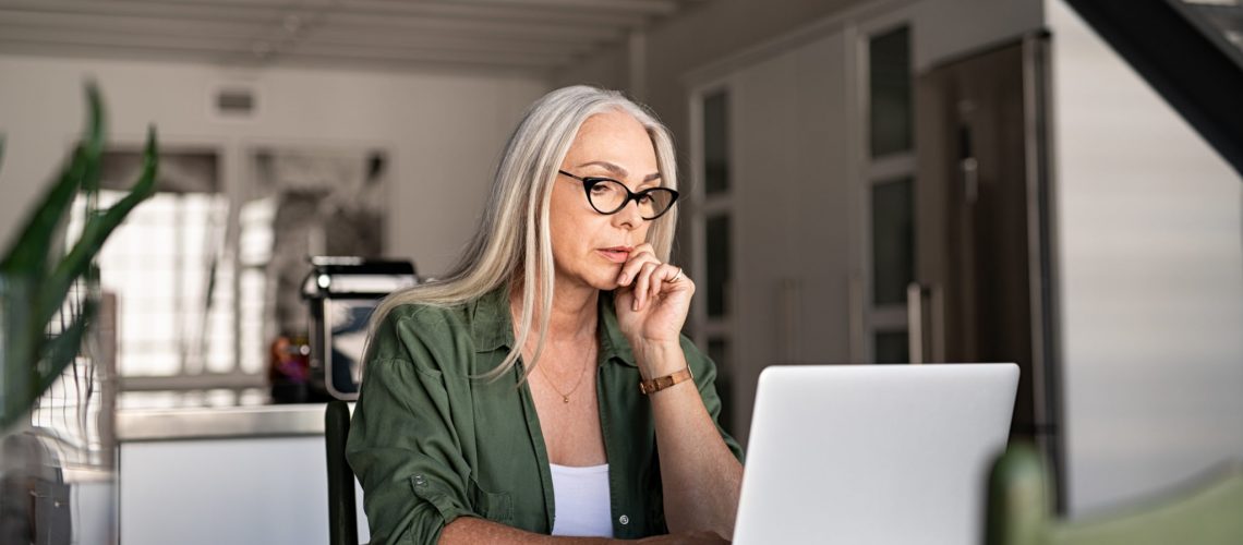 Focused old woman with white hair at home using laptop. Senior stylish entrepreneur with notebook and pen wearing eyeglasses working on computer at home. Serious woman analyzing and managing domestic bills and home finance.